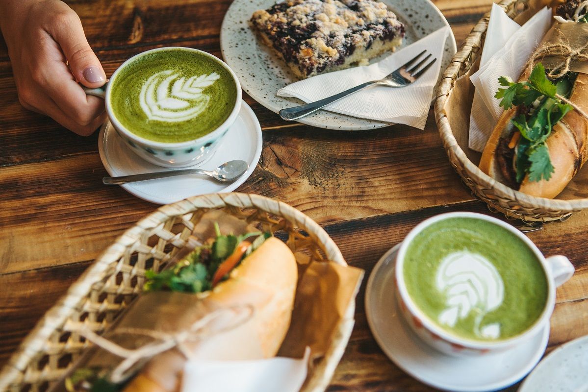 Top view – The girl sitting in cafe and holding mug with green tea with milk next to piece of sweet pie and two sandwiches with vegetables.