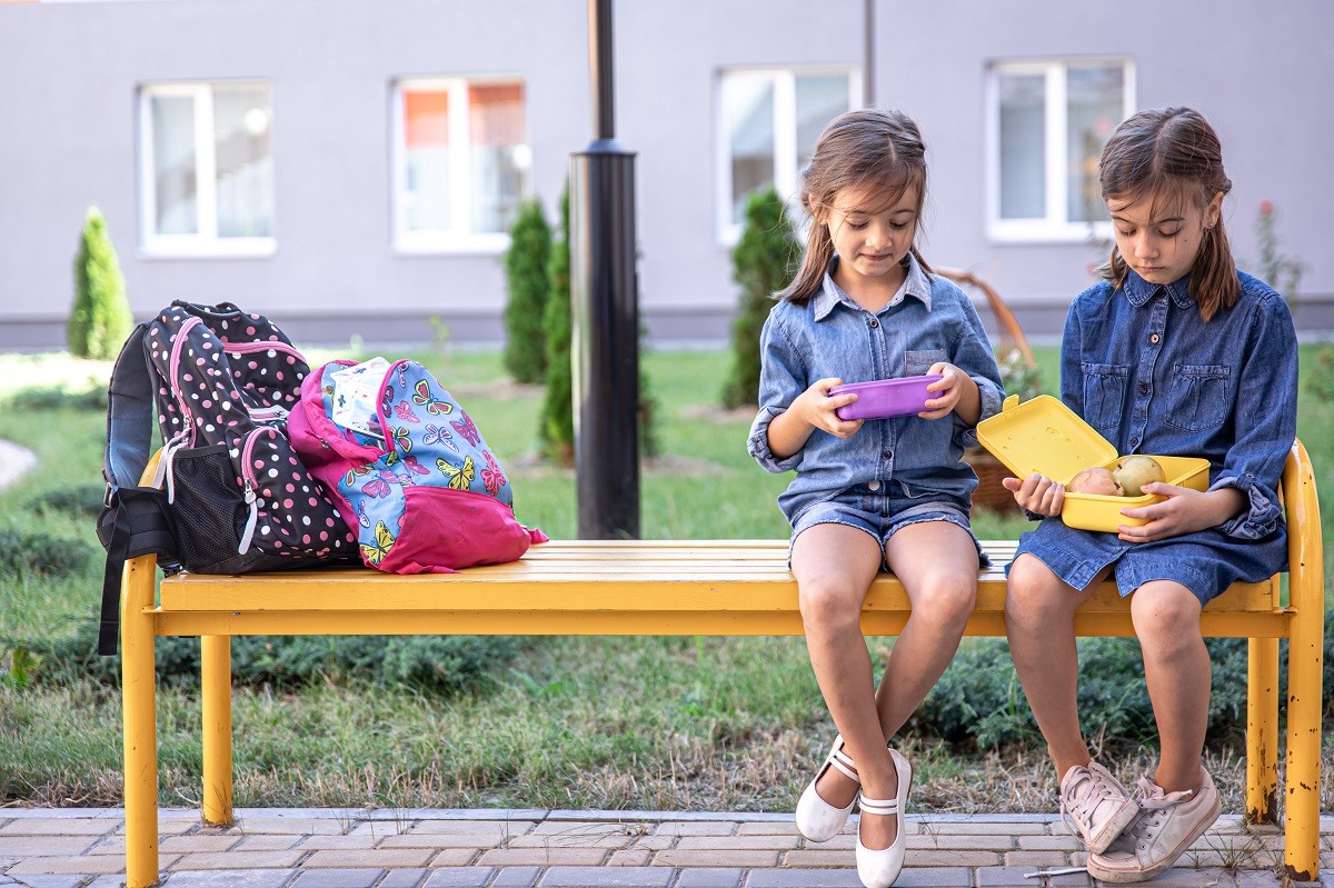 Little school girls sitting on bench in school yard and eating from lunch boxes.