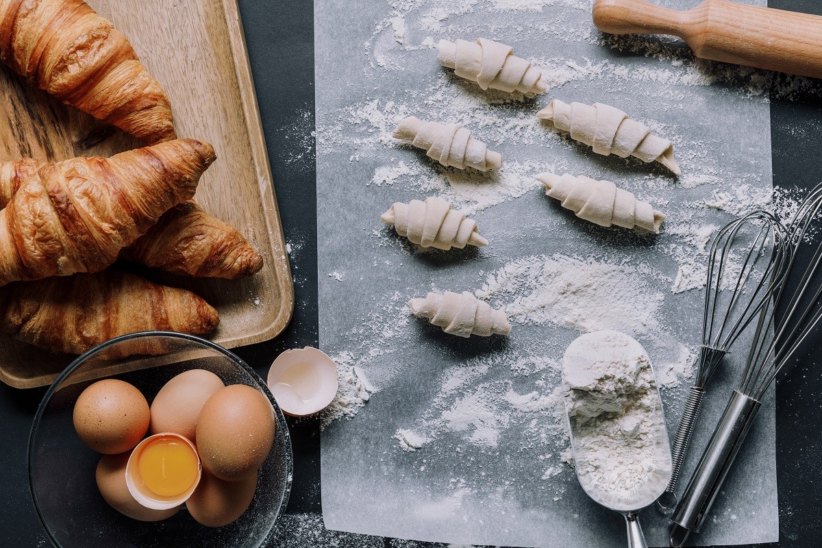 top view of rolling pin, whisks, dough for croissants on baking paper covered by flour