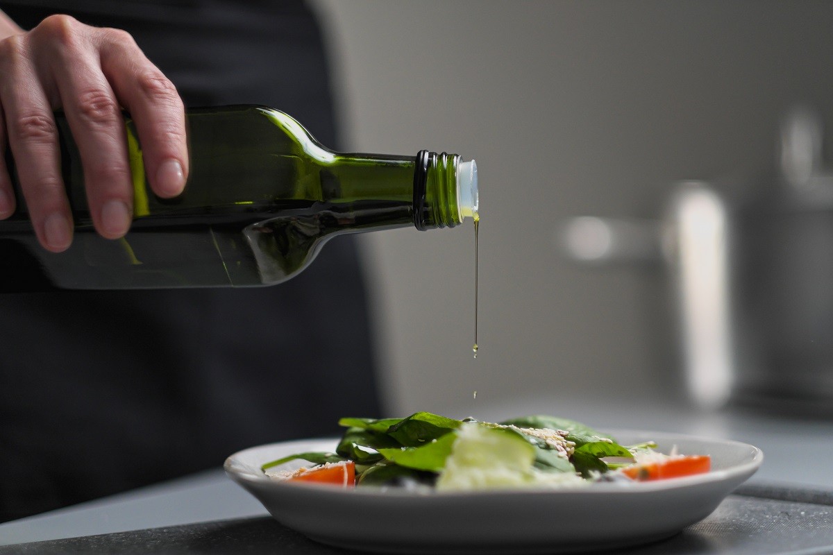 A female chef in a white uniform and a black apron in the restaurant kitchen. Cooking. The cook pours olive oil from a green bottle.