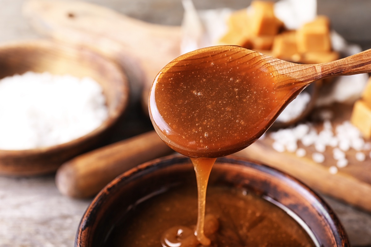 Spoon with tasty caramel sauce over bowl, closeup