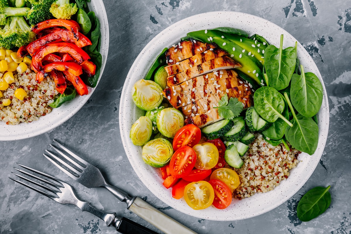 Healthy buddha bowl lunch with grilled chicken, quinoa, spinach, avocado, brussels sprouts, tomatoes, cucumbers  on dark grey background.