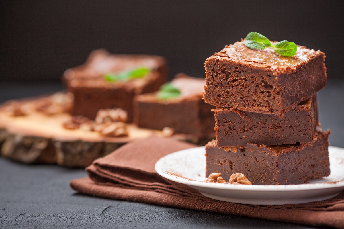 Chocolate brownie square pieces in stack on white plate with walnuts, decorated with mint leaves and cocoa on black background. Delicious dessert. Dark mood. Close up photography. Selective focus